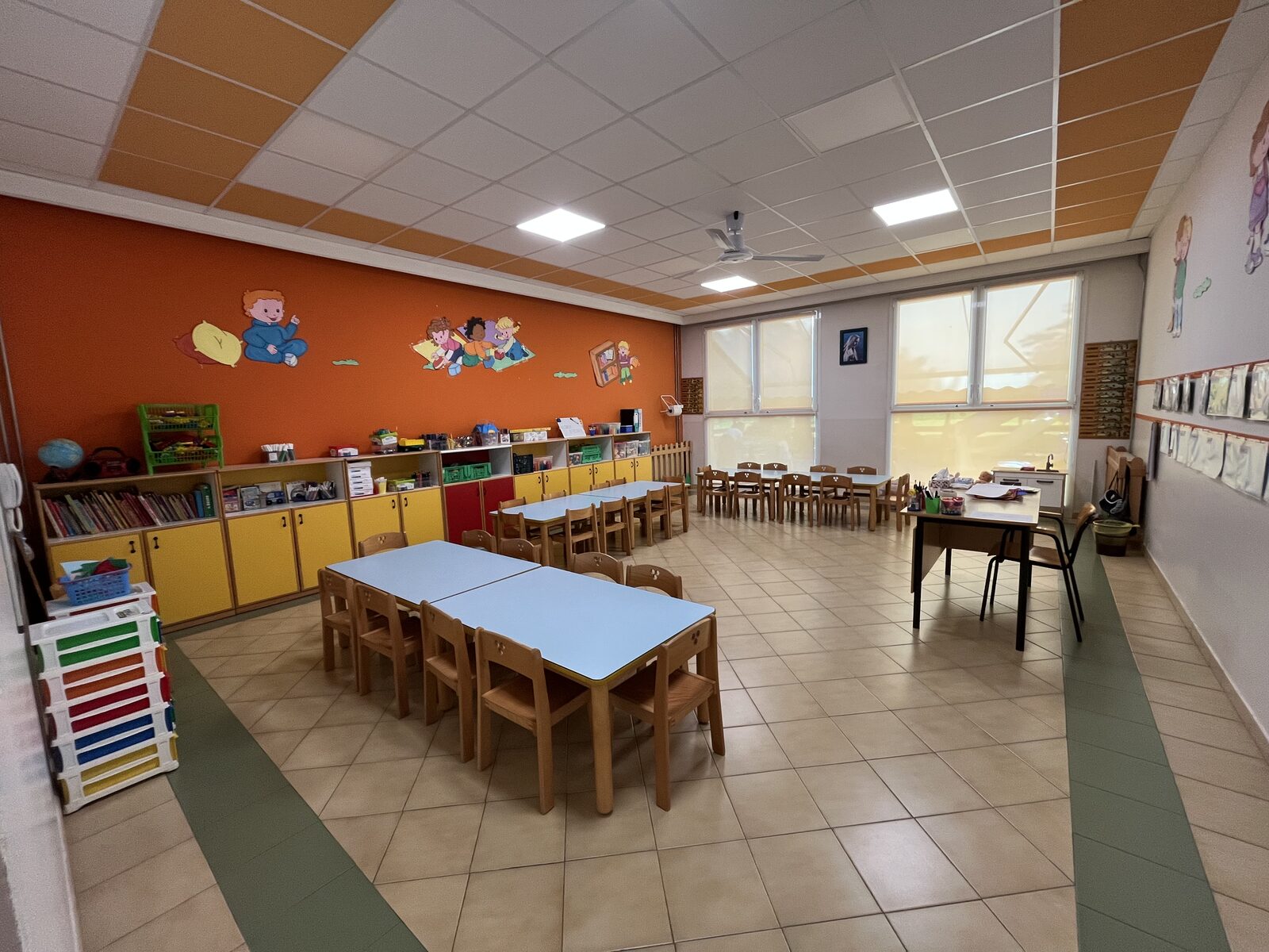 Un'aula luminosa della scuola con i tavolini azzurri, le sedioline di legno e la parete arancione decorata