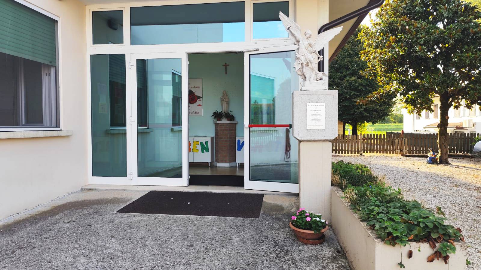 L'ingresso della scuola visto dall'esterno, con le porte a vetri, la statua dell'angelo sul pilastro e la scritta di benvenuto