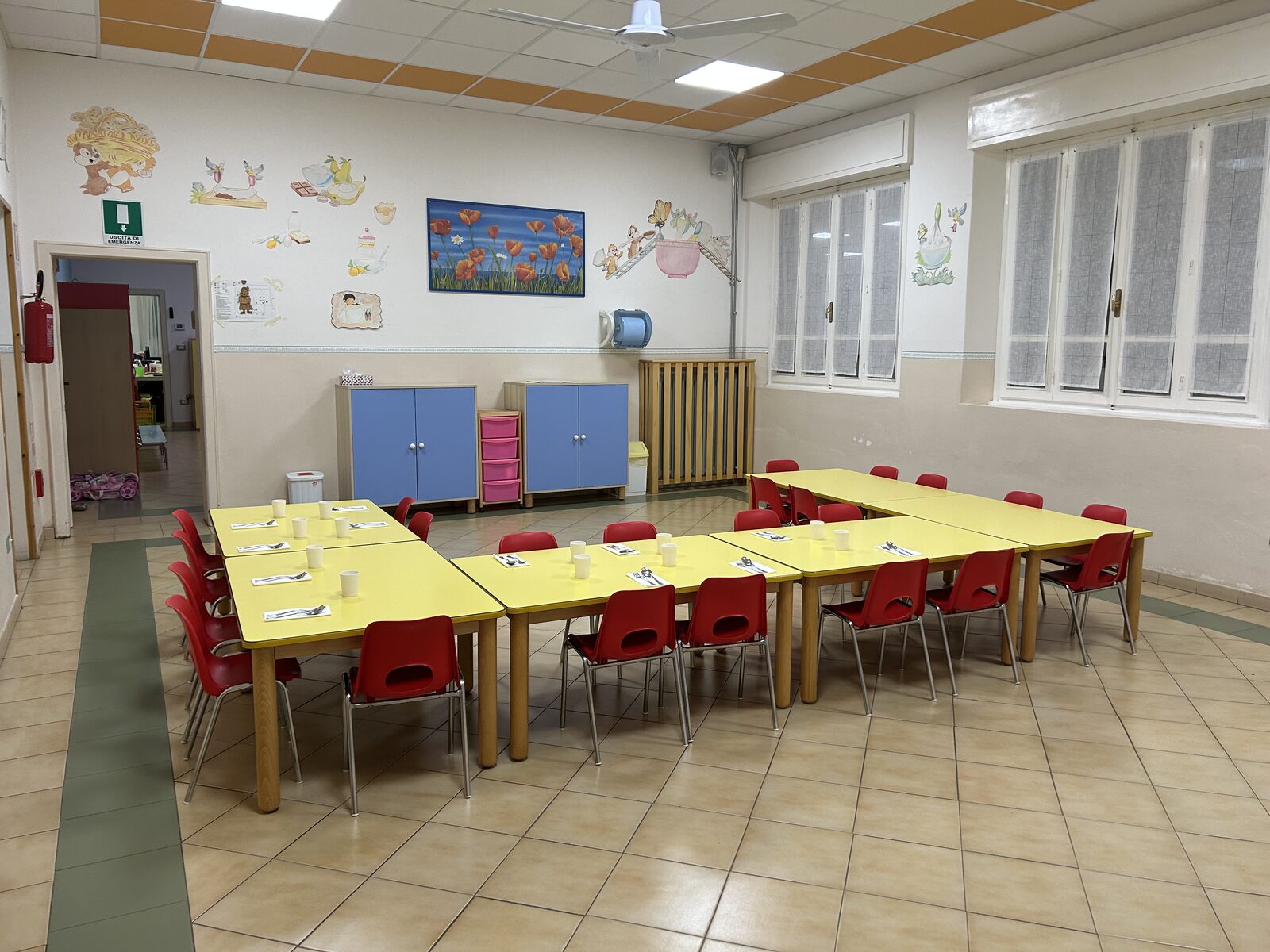 La sala da pranzo con i tavoli gialli apparecchiati, le seggioline rosse, gli armadietti azzurri e le decorazioni colorate alle pareti